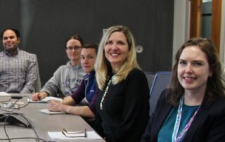 Jean Smart from Clinical Trials BC sitting at a table, surrounded by learners. They are all smiling with notebooks in front of them.