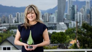 Stephania Manusha dressed in formal wear, holding a green glass trophy for the Clinical Trials BC awards. They are standing on a balcony in front of the Vancouver skyline.