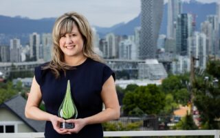 Stephania Manusha dressed in formal wear, holding a green glass trophy for the Clinical Trials BC awards. They are standing on a balcony in front of the Vancouver skyline.