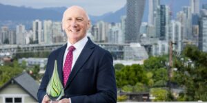Dr. Paul Anthony Keown dressed in formal wear, holding a green glass trophy for the Clinical Trials BC awards. They are standing on a balcony in front of the Vancouver skyline.