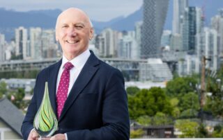 Dr. Paul Anthony Keown dressed in formal wear, holding a green glass trophy for the Clinical Trials BC awards. They are standing on a balcony in front of the Vancouver skyline.