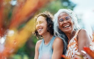 An adult daughter and her senior mother laughing outdoors, holding each other close. Behind and in front of them are green and orange foliage.