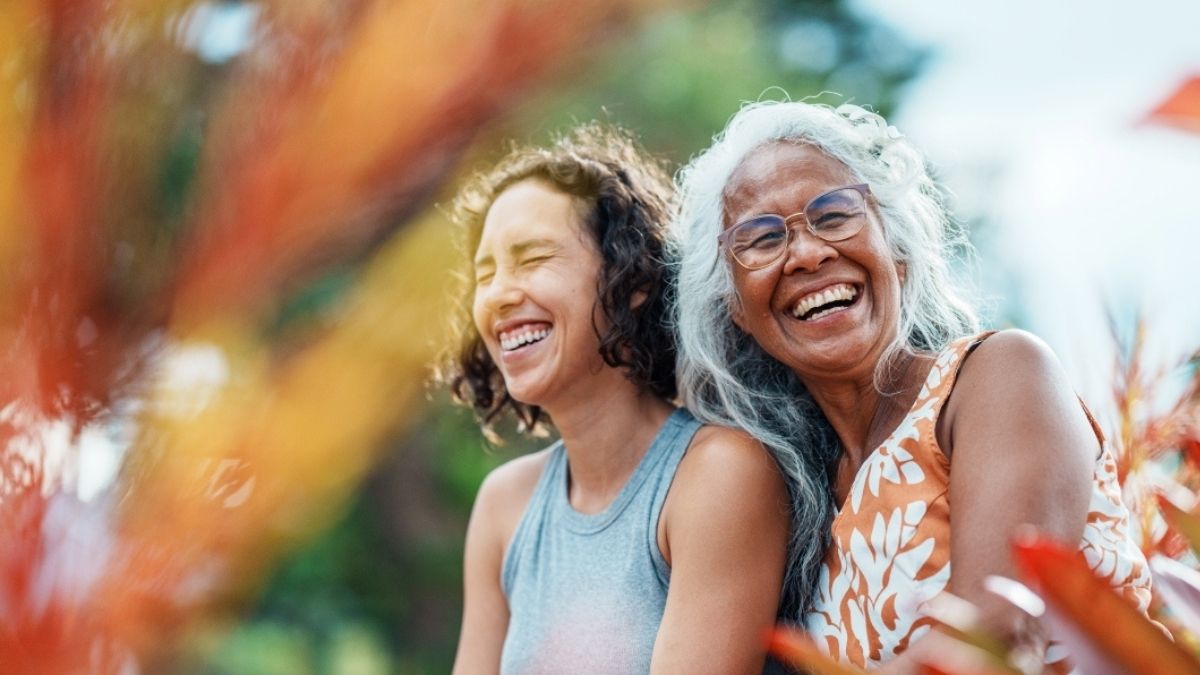 An adult daughter and her senior mother laughing outdoors, holding each other close. Behind and in front of them are green and orange foliage.