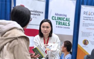 A REACH BC team member speaks to an attendee at The Wellness Show 2026. There are three banners behind the team member: Providence Research, Clinical Trials British Columbia, and REACH BC.