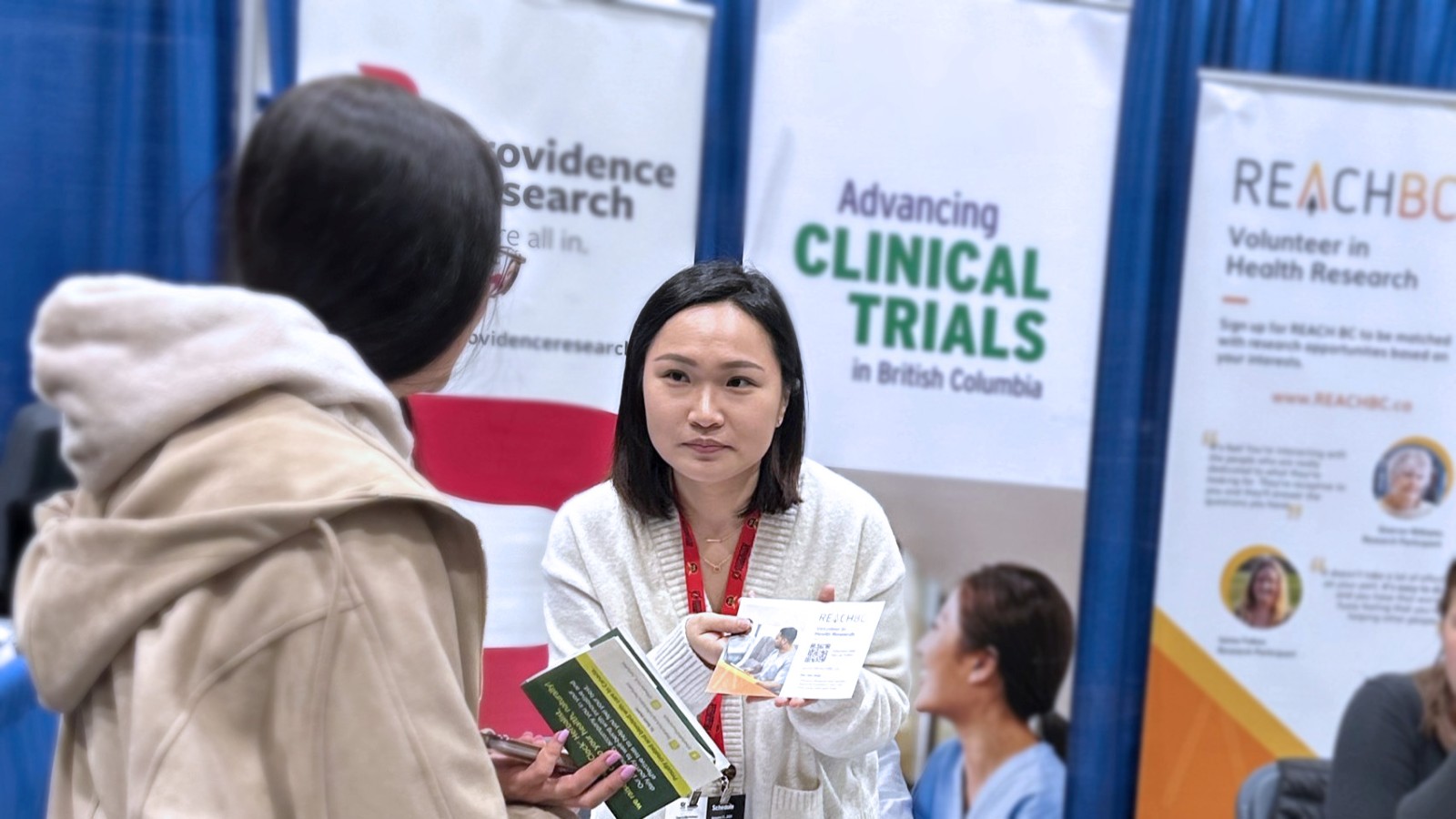 A REACH BC team member speaks to an attendee at The Wellness Show 2026. There are three banners behind the team member: Providence Research, Clinical Trials British Columbia, and REACH BC.