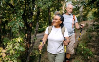 Three middle age to senior friends hiking together in the woods. They are walking single file and holding hiking poles, looking around them in delight.