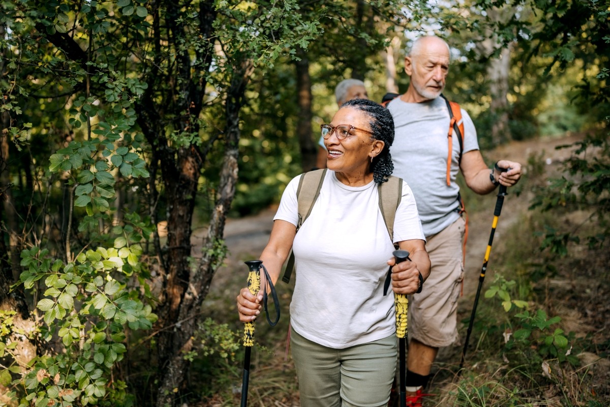 Three-senior-friends-hiking-together_iStock-2250105047 Three middle age to senior friends hiking together in the woods. They are walking single file and holding hiking poles, looking around them in delight.